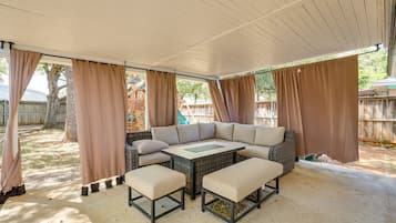 A covered patio with a sectional sofa and two matching ottomans, a small table, and a view of a wooden fence and trees.