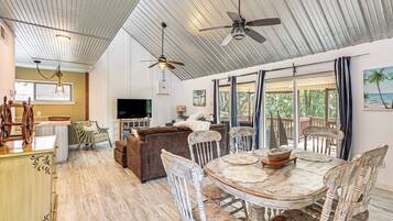 A spacious living area with a high ceiling, a dining table with chairs, a television, and a view of the outdoors through large windows.