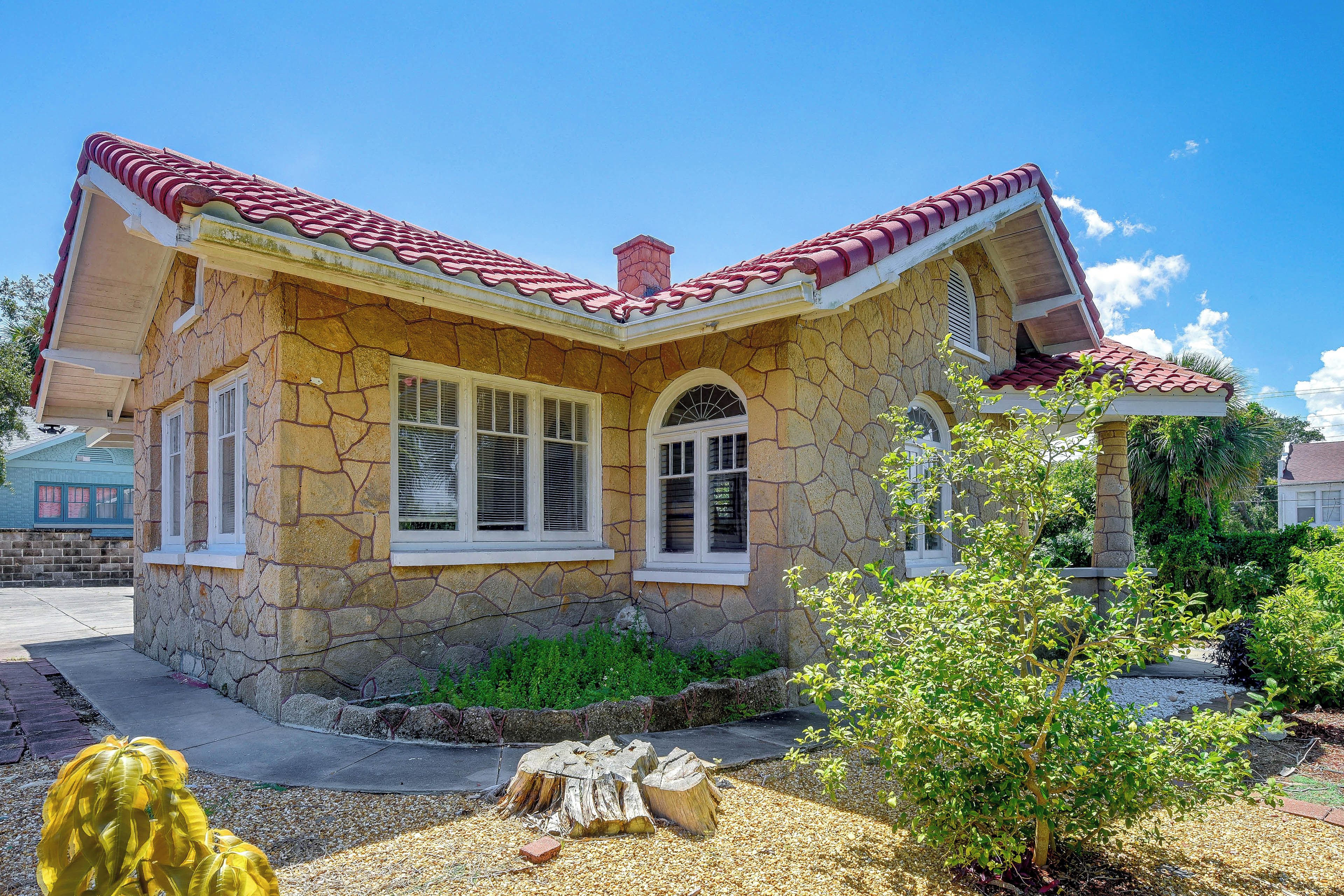 Uma casa de pedra com telhado de telhas vermelhas, duas janelas com persianas e um pequeno jardim com uma árvore e um toco de madeira.