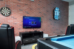 Ein Zimmer mit Backsteinwand, Dartscheibe, Flachbildfernseher, Gaming-Stühlen und einem Schild für eine Veranstaltung.