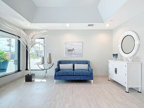 A modern living room with a blue sofa, a white cabinet, and a large window. - Beachfront Getaway at Caper Beach Club #803 | Private Chiller/Heated Pool (Fort Myers Beach)