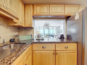 A kitchen with wooden cabinets, granite countertops, a stainless steel sink, and a refrigerator. - Beachfront Getaway at Caper Beach Club #803 | Private Chiller/Heated Pool (Fort Myers Beach)