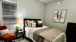 Ein Schlafzimmer mit Bett, Nachttisch, Lampe, Stuhl und Kunstwerk an der Wand.
