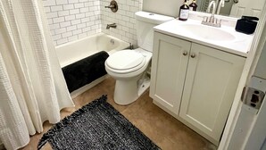 Ein Badezimmer mit weiß gefliesten Wänden, einer Badewanne, einer Toilette, einem Waschbecken mit Spiegel und einem schwarz-weißen Teppich auf dem Boden.
