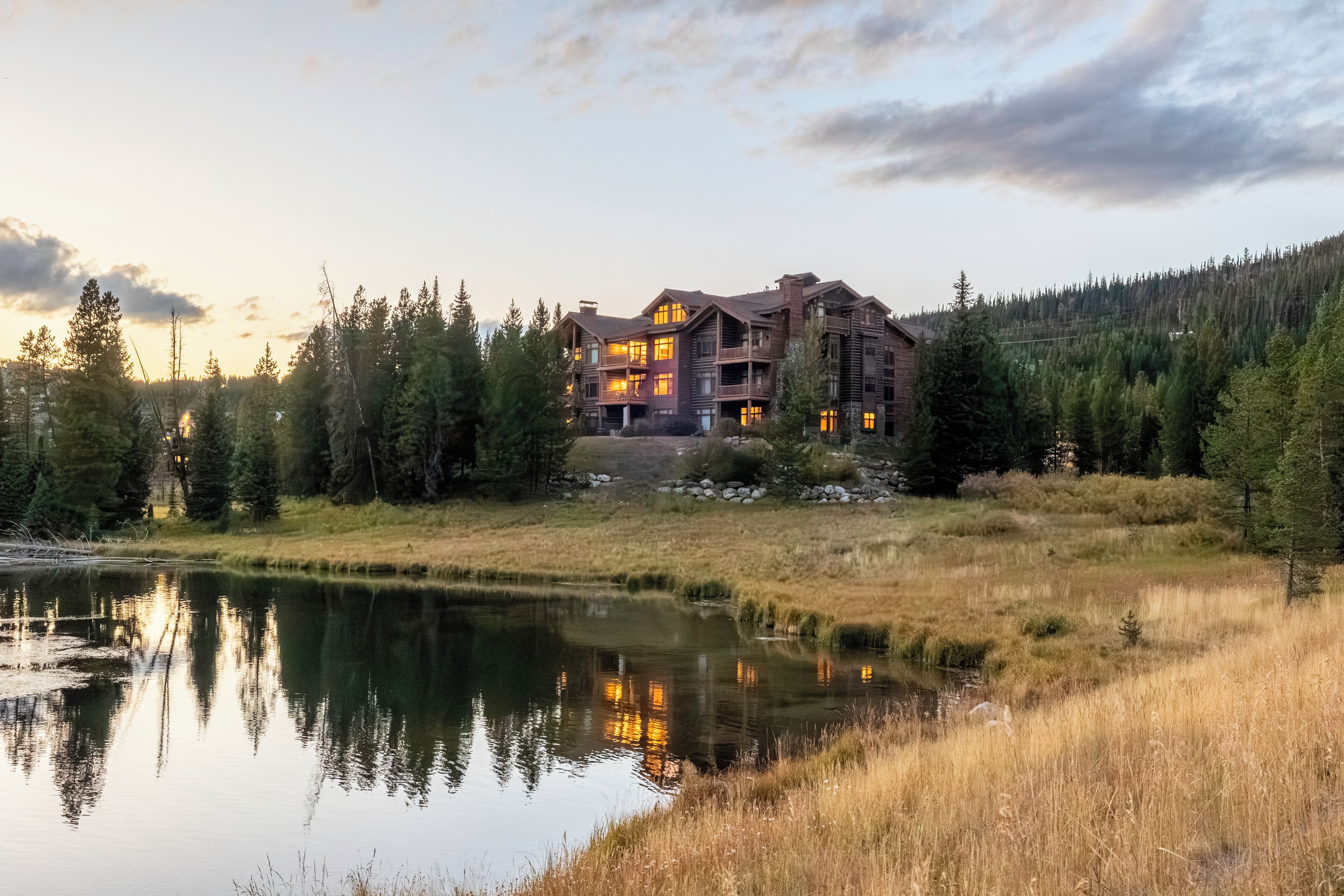 A large, multi-story log cabin with a reflective pond in front, surrounded by dense forest.