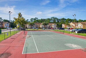 A tennis court with a net, surrounded by a fence, and adjacent to a parking area with multiple cars. - Whimsical Wonders Sweetwater Club Near Disney (Kissimmee)