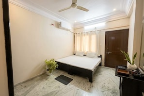 A bedroom with a bed, a ceiling fan, an air conditioning unit, a potted plant, a desk with a chair, and a door. - The Shyam Haven (UJJAIN)