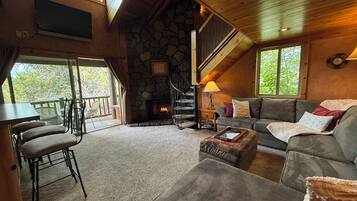 A cozy living room with a stone fireplace, a staircase, a sofa, and a dining area with chairs.