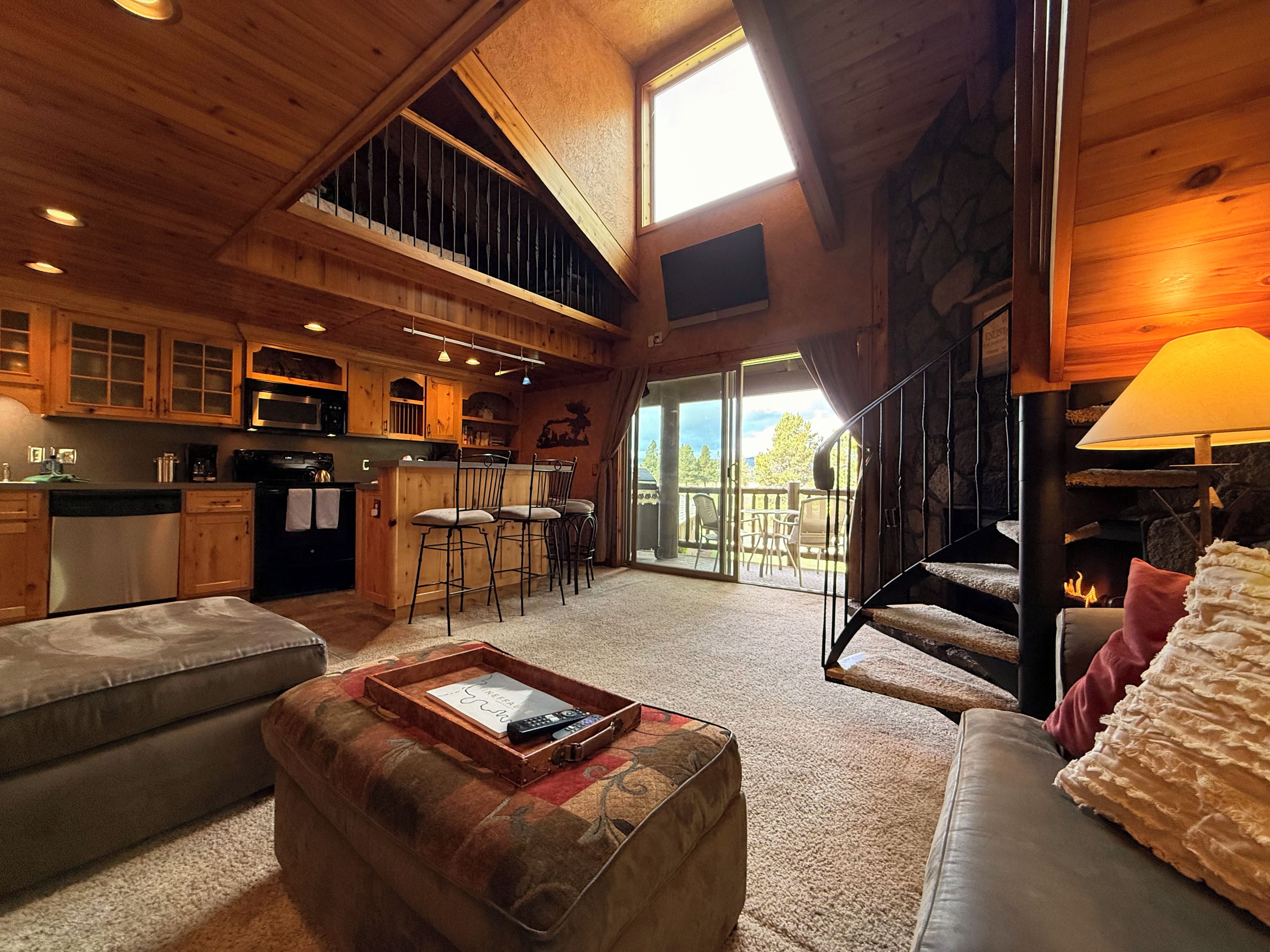 A spacious living area with a high ceiling, wooden beams, a fireplace, and a large window overlooking a deck.