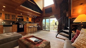 A spacious living area with a high ceiling, wooden beams, a fireplace, and a large window overlooking a deck.