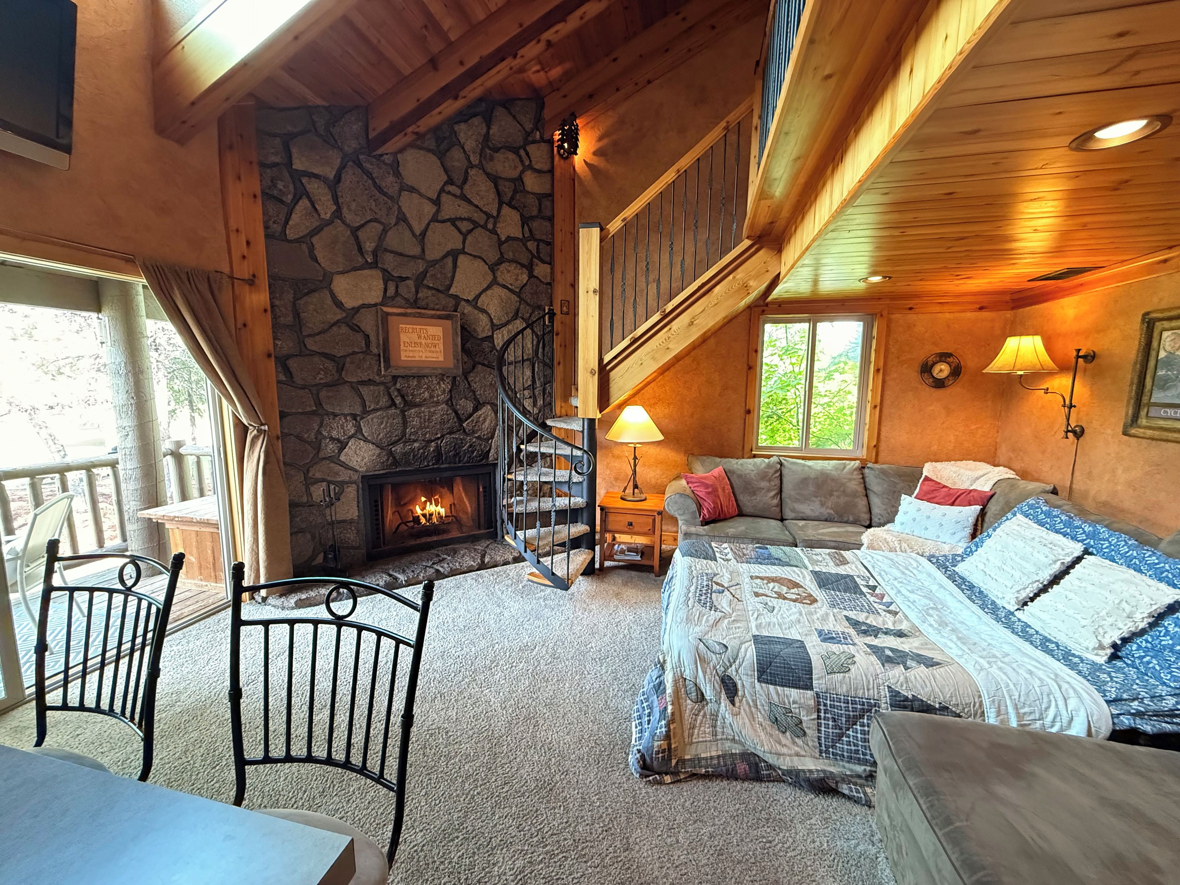 A cozy cabin interior with a stone fireplace, a wooden staircase, a sofa, and a bed with a quilt.