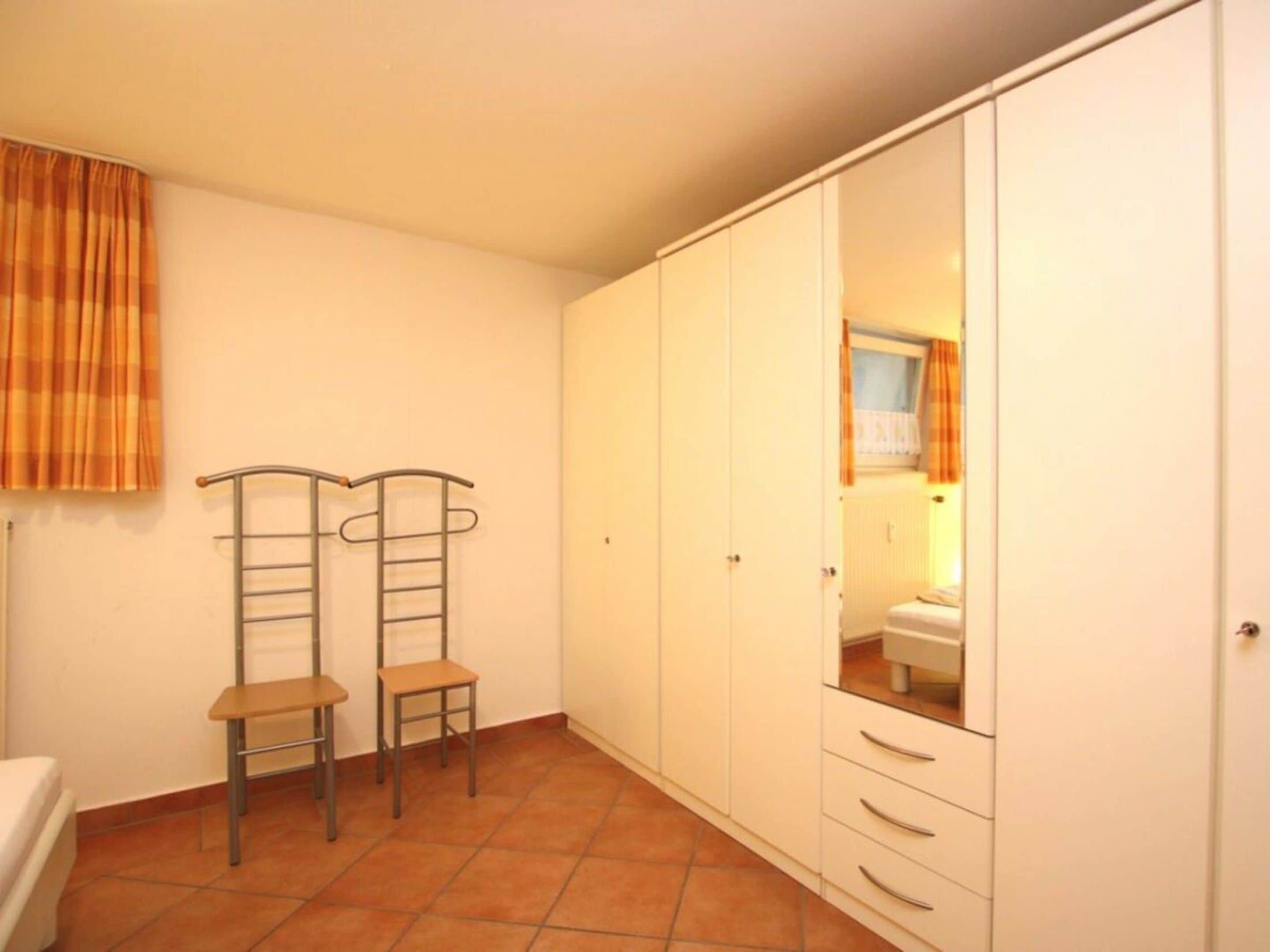 Een kamer met een kledingkast, twee stoelen, een klein tafeltje en een raam met gordijnen.