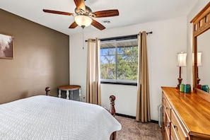 Un dormitorio con cama, ventilador de techo, ventana con cortinas, mesita de noche, cómoda y lámpara.