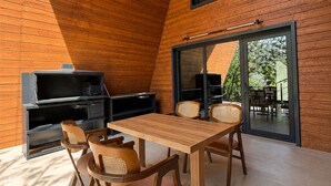 A modern interior with a wooden sloped ceiling, a dining area with a table and chairs, a built-in entertainment unit, and a large sliding glass door leading to an outdoor area. - Oxygen garden (Yangiabad)