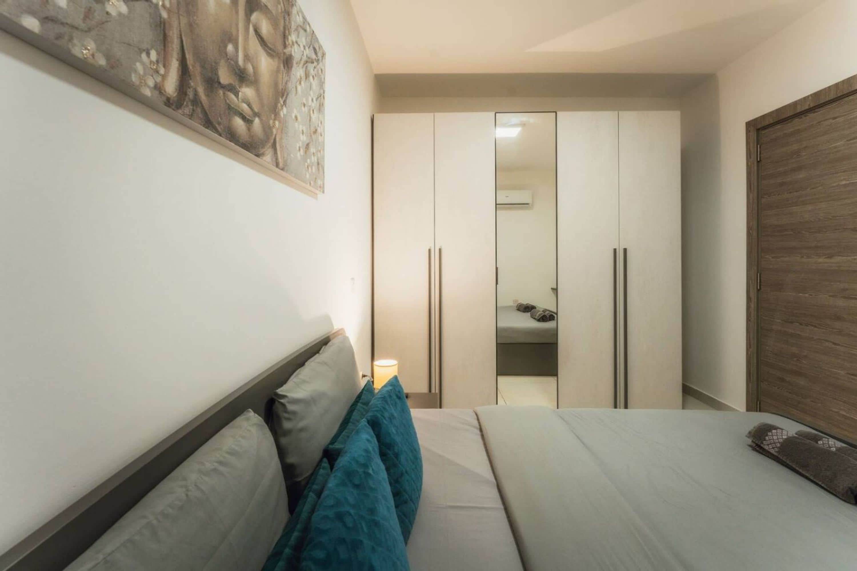 Een slaapkamer met een bed, kussens, een nachtkastje, een kledingkast en een spiegel.