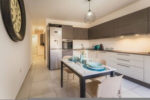 A modern kitchen with a dining area, featuring a clock on the wall, a refrigerator, an oven, and a pendant light. - Zen Apartment in the Heart of Malta (Msida)