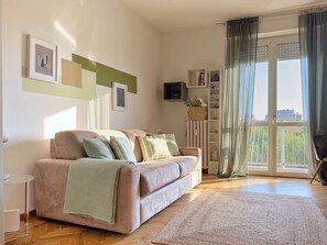 A modern living room with a sofa, a coffee table, a TV, and a view of the outdoors through a large window. - The Washington Studio Apartment - with balcony (Milano)