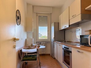 A compact kitchen with a dining area, a window with blinds, and a view of buildings outside. - The Washington Studio Apartment - with balcony (Milano)