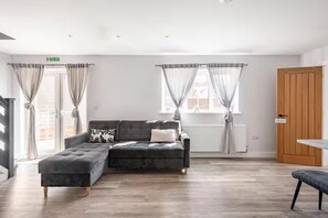 Ein modernes Wohnzimmer mit einer Eckcouch, einem kleinen Tisch und einer Bank. Es gibt zwei Fenster mit Vorhängen, eine Heizung und eine Holztür.