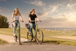Two individuals riding bicycles on a paved path with a grassy field and a clear sky in the background. - Küstenherz Strandhotel (Otterndorf)