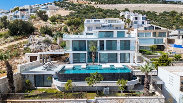 Um edifício moderno de vários andares com piscina no terraço, rodeado por um morro rochoso e vegetação exuberante.