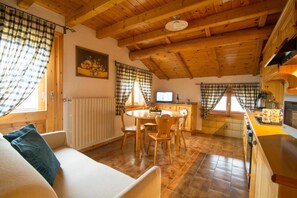 Een knus vertrek met houten plafond, een eethoek met tafel en stoelen, een keuken met fornuis en spoelbak, en een raam met gordijnen.
