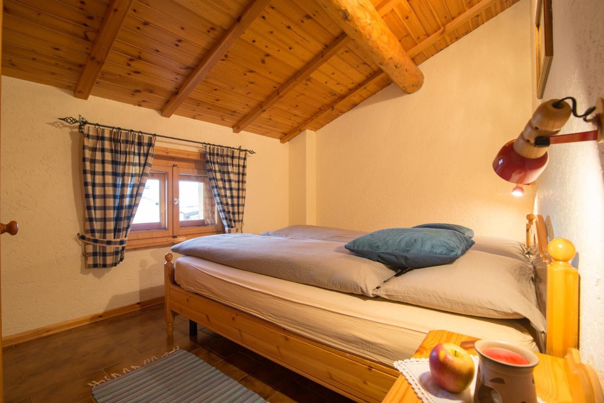 Una camera da letto con un letto di legno, una finestra con tende a quadri, una lampada da comodino e un tavolino con frutta e una candela.