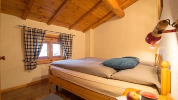 Una camera da letto con un letto di legno, una finestra con tende a quadri, una lampada da comodino e un tavolino con frutta e una candela.