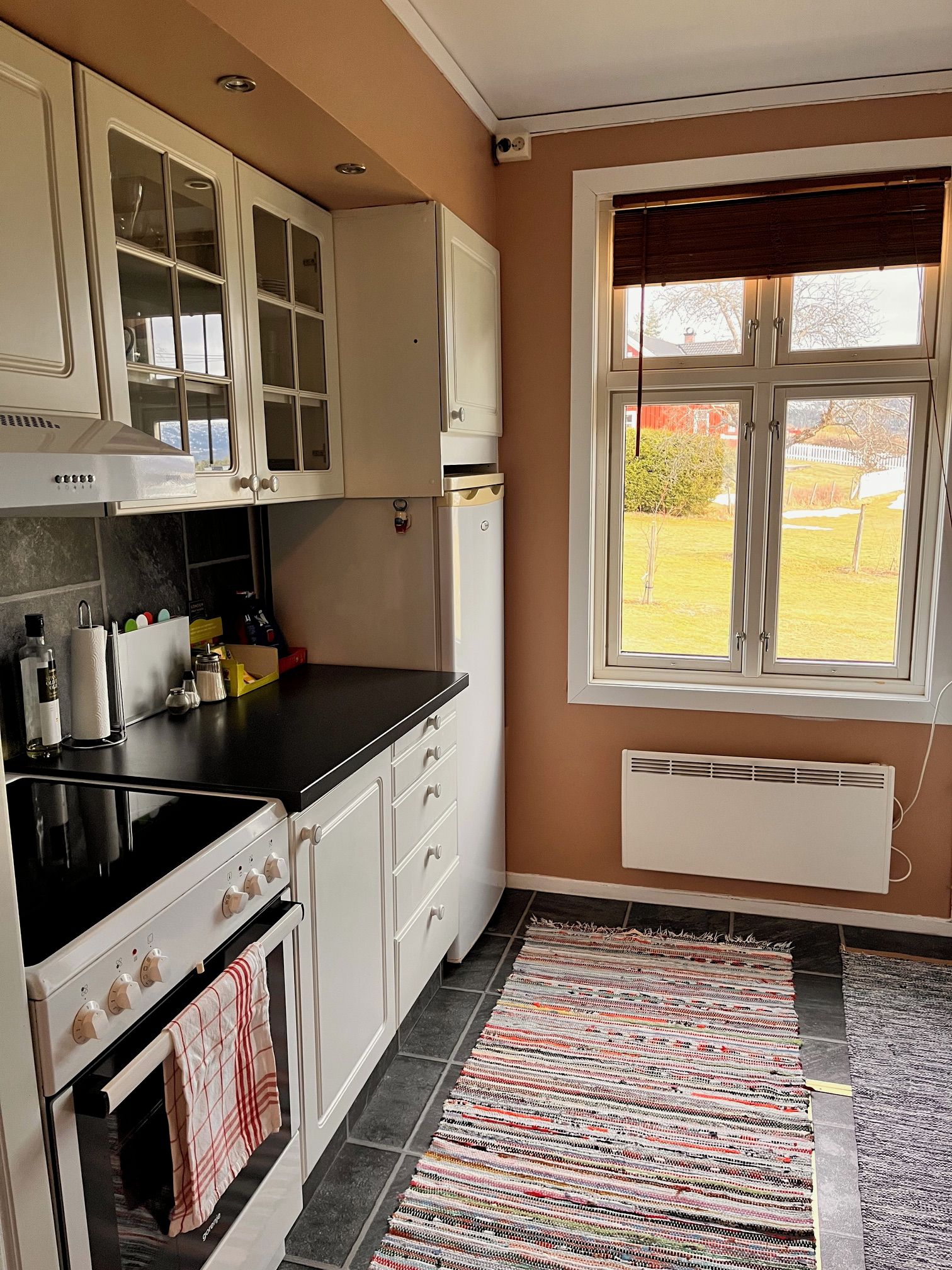 Une cuisine avec des meubles blancs, un plan de travail noir, une cuisinière et une fenêtre avec des stores.