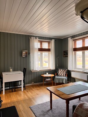 Une pièce avec une table en bois, une chaise, une petite table ronde avec une lampe et une fenêtre avec des rideaux.