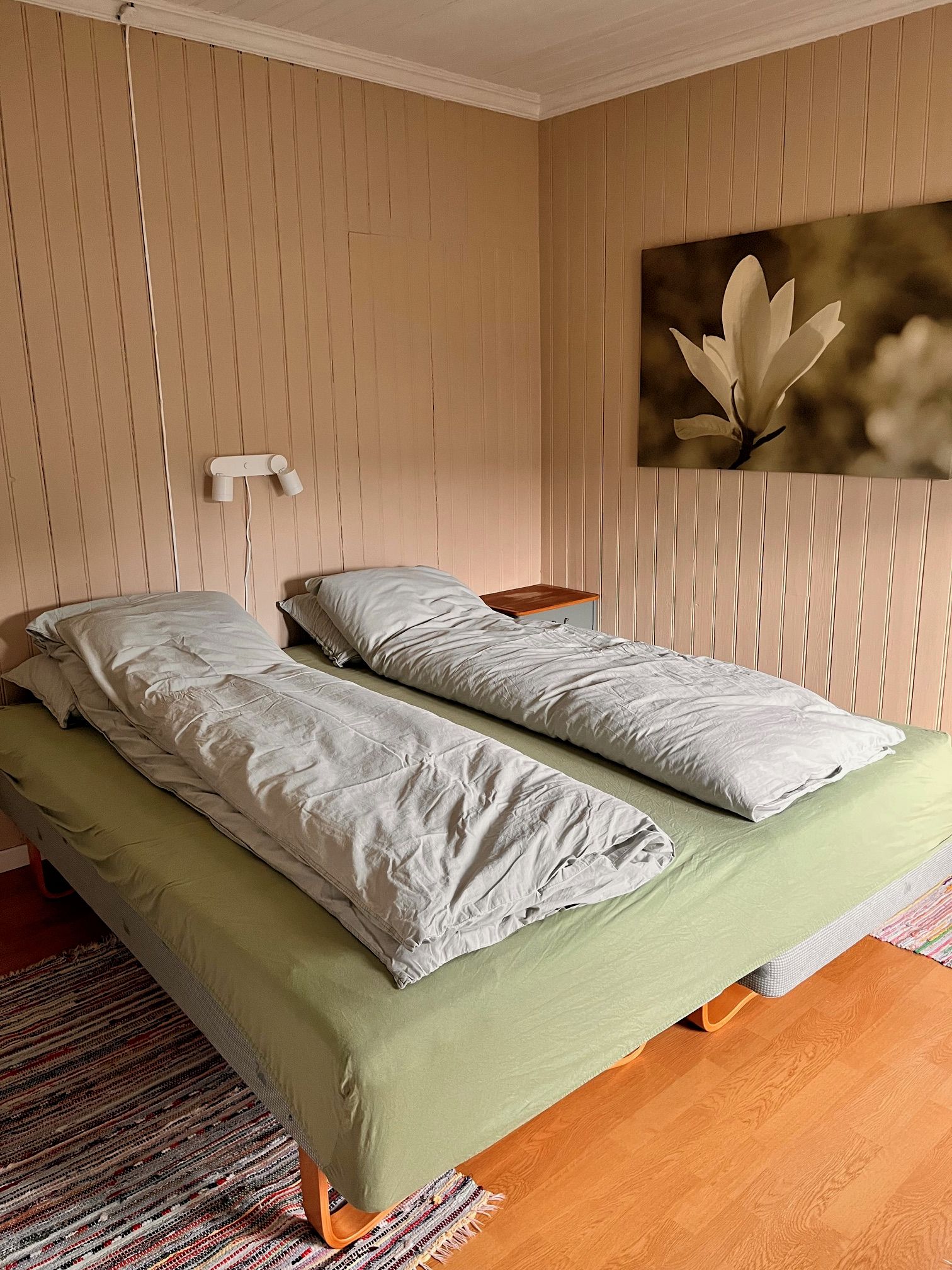 Un lit double avec une couvre-lit verte et deux oreillers, une table de chevet en bois, une lampe fixée au mur et un tableau représentant une fleur.