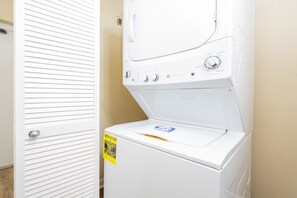 A front-loading white washer and dryer unit with a control panel, a yellow energy guide sticker, and a door with horizontal slats. - Phoenix Vi 61407 1 Bedroom Condo (Orange Beach)