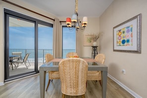 A dining area with a glass table, wicker chairs, a chandelier, and a view of the ocean. - Phoenix Vi 61407 1 Bedroom Condo (Orange Beach)