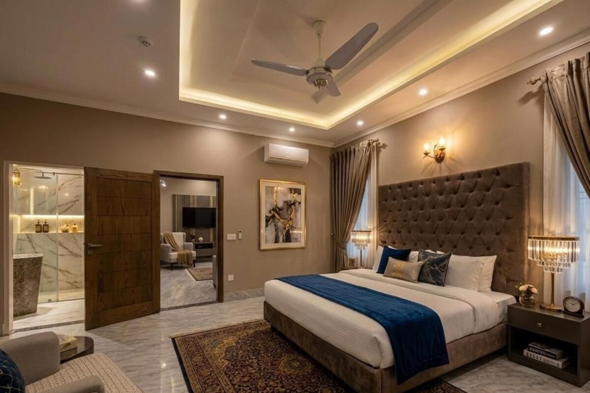 Ein Schlafzimmer mit einem großen Bett, einem Deckenventilator, einem Nachttisch mit Lampe und Blick ins Badezimmer.