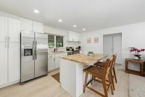 A modern kitchen with white cabinetry, a stainless steel refrigerator, a wooden island, and a dining area with chairs. - Big Island Kahaluu Surf Boutique #5 2 Bedroom Apts (Kailua-Kona)