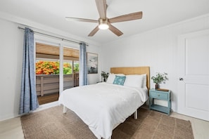 A bedroom with a bed, a nightstand, a ceiling fan, and a view of a balcony with flowers. - Big Island Kahaluu Surf Boutique #5 2 Bedroom Apts (Kailua-Kona)