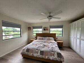 Une chambre à coucher comprenant un lit, un ventilateur de plafond, deux tables de chevet et une fenêtre donnant sur un paysage verdoyant.