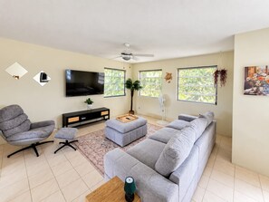A modern living room with a grey sofa, a TV, a ceiling fan, and a large window with a view of greenery. - Modern 2 bed 2 bath home away from home perfectly located for visitors (Bridgetown)