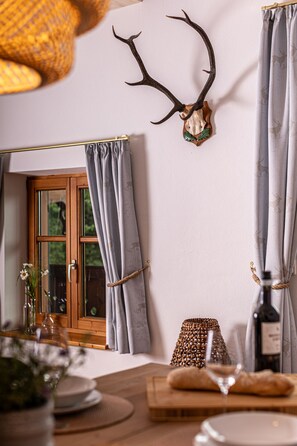 Une pièce avec une table en bois, une bouteille de vin, un panier en osier et des rideaux attachés avec une corde.