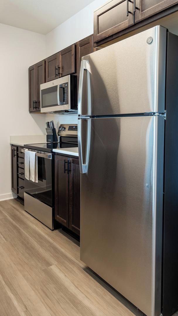 A modern kitchen with stainless steel appliances, dark wooden cabinets, and a light wood floor.