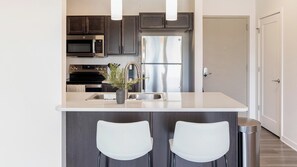 A modern kitchen with dark cabinets, stainless steel appliances, a white countertop, and two white bar stools.