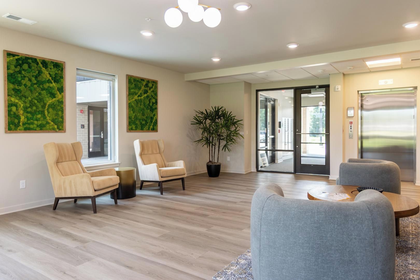 A modern waiting area with a wooden floor, two beige armchairs, a grey sofa, a potted plant, and a glass door leading outside.