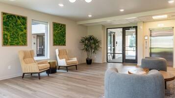 A modern waiting area with a wooden floor, two beige armchairs, a grey sofa, a potted plant, and a glass door leading outside.