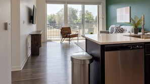 A modern kitchen with a dishwasher, a dining area with a chair, and a view through a glass door to an outdoor balcony.