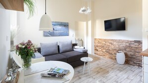 A modern living room with a grey sofa, a round dining table with a vase of flowers, a flat-screen TV mounted on the wall, and a wooden accent wall. - Vrbo Property (Norderney)