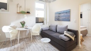 A modern living room with a grey sofa, a white chair, a small round table, and a large window with blinds. - Vrbo Property (Norderney)