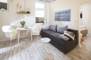 Ein modernes Wohnzimmer mit einem grauen Sofa, einem weißen Sessel, einem kleinen runden Tisch und einem großen Fenster mit Jalousien.