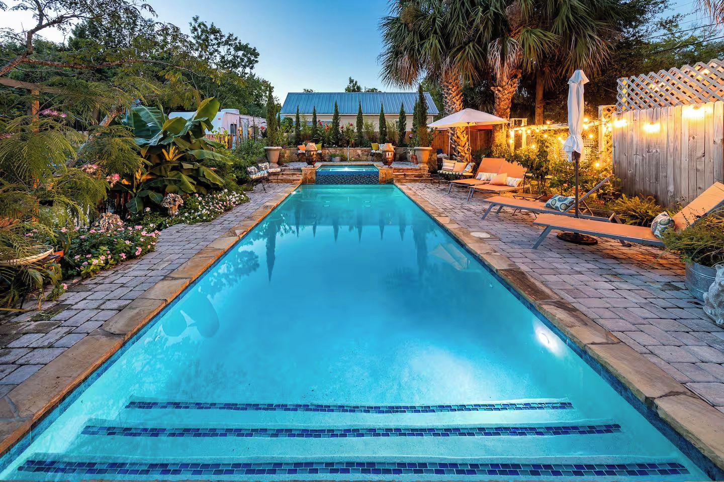 A rectangular swimming pool with a tiled bottom, surrounded by a brick walkway, outdoor furniture, and lush greenery.