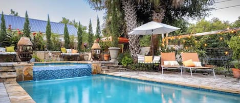 A swimming pool with lounge chairs and umbrellas, surrounded by potted plants and a pergola with string lights.
