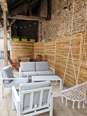 An outdoor seating area with a wooden table and chairs, a hanging chair, and a wall with a wooden panel design. - Vrbo Property (Châteauvillain)